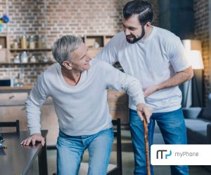 Zdjęcie dwóch mężczyzn, z którym młodszy pomaga starszemu wstać z krzesła. Starszy mężczyzna jedną rękę opartą ma na lasce, a drugą o blat stolu. Uśmiecha się do młodszego mężczyzny. W tle lekko rozmyte wyposażenie kuchenne.