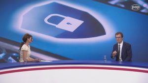 Zdjęcie kobiety i mężczyzny w studiu telewizyjnym, siedzących przy okrągłym stole, nad którym widać grafikę przedstawiającą zamkniętą kłódkę w swoistej tarczy będącej z kolei na ekranie telefonicznym.